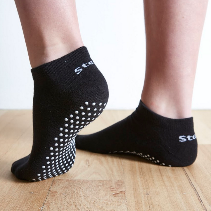 Image of Black Stealth Movement Pilates Grip Socks. The image is taken from the back, with the left foot showing the white grips underneath. The right foot is angled to show the inside of the foot, showing a small part of the white grips underneath. The back of both socks say "Stealth.”