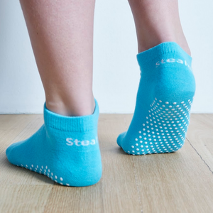 Image of Aqua Stealth Movement Pilates Grip Socks. The image is taken from the back, with the right foot fully showing the white grips underneath. The left foot is angled to show the heel of the foot, a small part of the white grips can be seen on the outside of the foot. The back of the socks say "Stealth.”