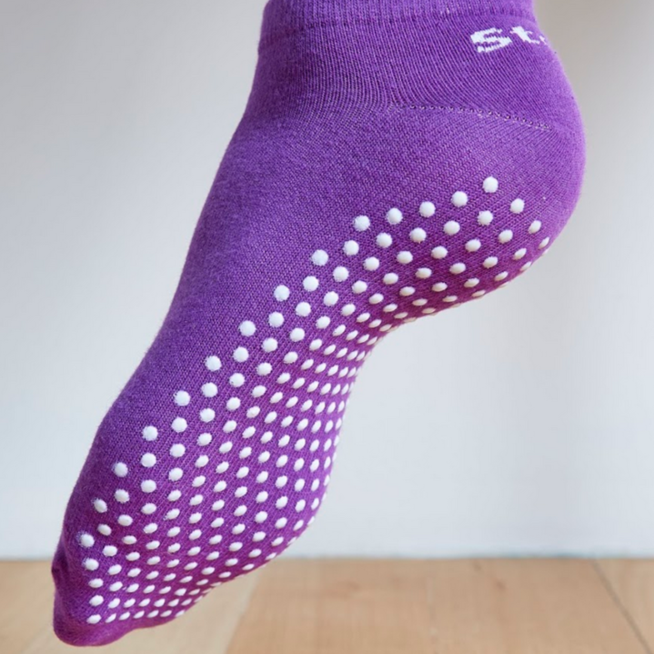 Image of a Purple Stealth Movement Pilates Grip Sock. The image is taken from the back, with the left foot showing the white grips underneath. The back of the sock says "Stealth.”