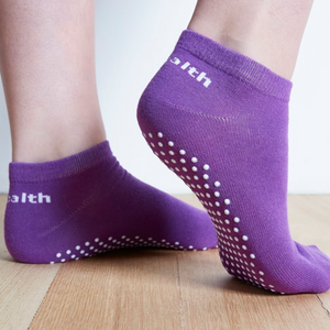 Image of Purple Stealth Movement Pilates Grip Socks. The image is taken from the back, with the right foot showing the white grips underneath. The left foot is angled to show the inside of the foot, showing a small part of the white grips underneath. The back of both socks say "Stealth.”