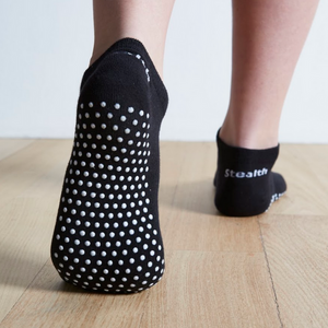 Image of Black Stealth Movement Pilates Grip Socks. The image is taken from the back, with the left foot fully showing the white grips underneath. The right foot is angled to show the heel of the foot, a small part of the white grips can be seen on the outside of the foot. The back of the socks say "Stealth.”