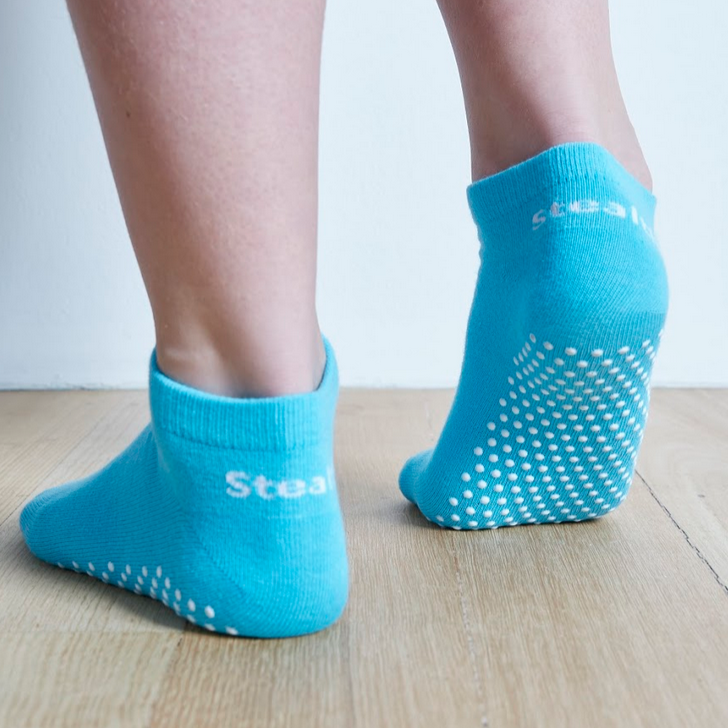 Image of Aqua Stealth Movement Pilates Grip Socks. The image is taken from the back, with the right foot fully showing the white grips underneath. The left foot is angled to show the heel of the foot, a small part of the white grips can be seen on the outside of the foot. The back of the socks say "Stealth.”