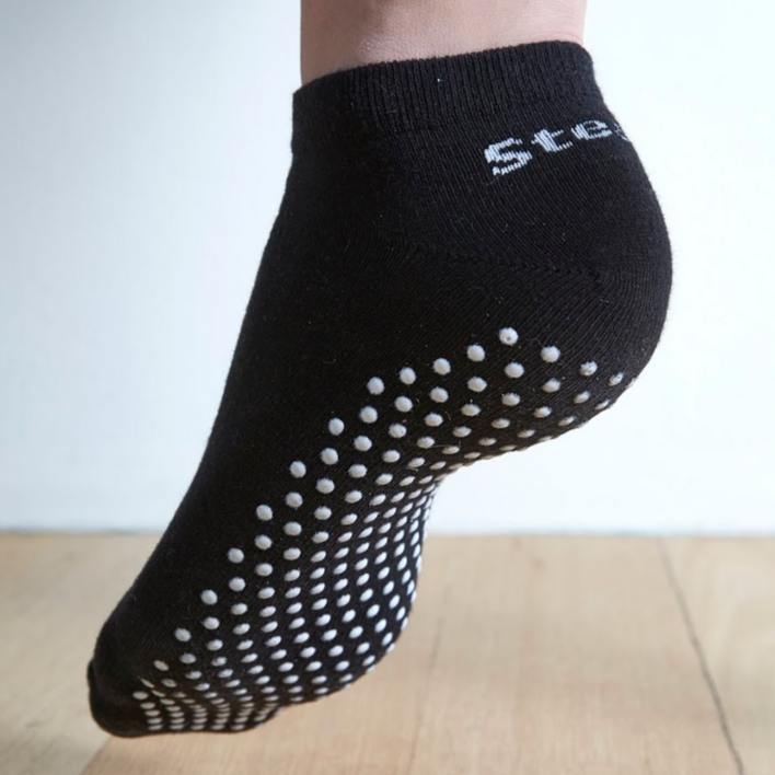 Image of a Black Stealth Movement Pilates Grip Sock. The image is taken from the back, with the left foot showing the white grips underneath. The back of the sock says "Stealth.”