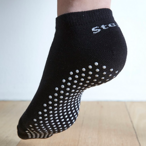 Image of a Black Stealth Movement Pilates Grip Sock. The image is taken from the back, with the left foot showing the white grips underneath. The back of the sock say "Stealth.”